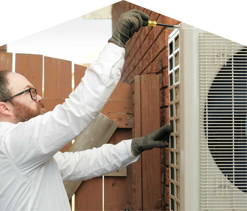 M&K service personnel working on an AC condenser, using a screwdriver to secure the unit’s panel outdoors beside a wooden fence. M&K service personnel working on an AC condenser, using a screwdriver to secure the unit's panel outdoors beside a wooden fence.