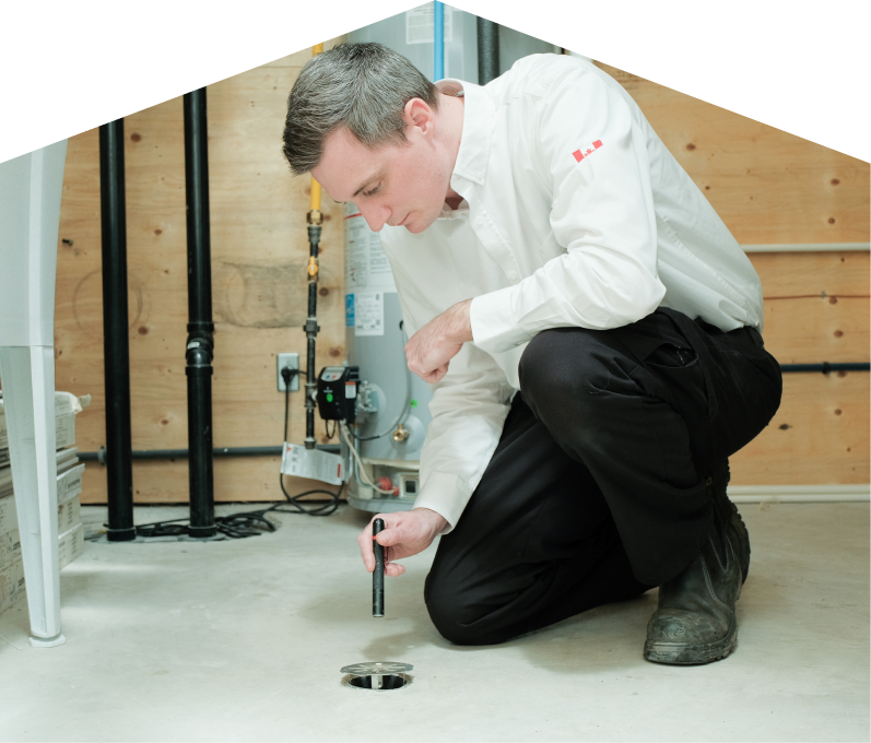 M&K plumbing specialist inspecting a basement floor drain with a flashlight during a drain cleaning service, in a utility room with visible pipes, water heater, and wood panel walls. M&K plumbing specialist inspecting a basement floor drain with a flashlight during a drain cleaning service, in a utility room with visible pipes, water heater, and wood panel walls.