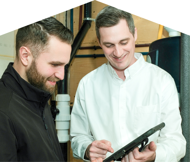 M&K specialist reviewing maintenance plan options with a smiling homeowner using a tablet, inside a utility room with visible piping. M&K specialist reviewing maintenance plan options with a smiling homeowner using a tablet, inside a utility room with visible piping.