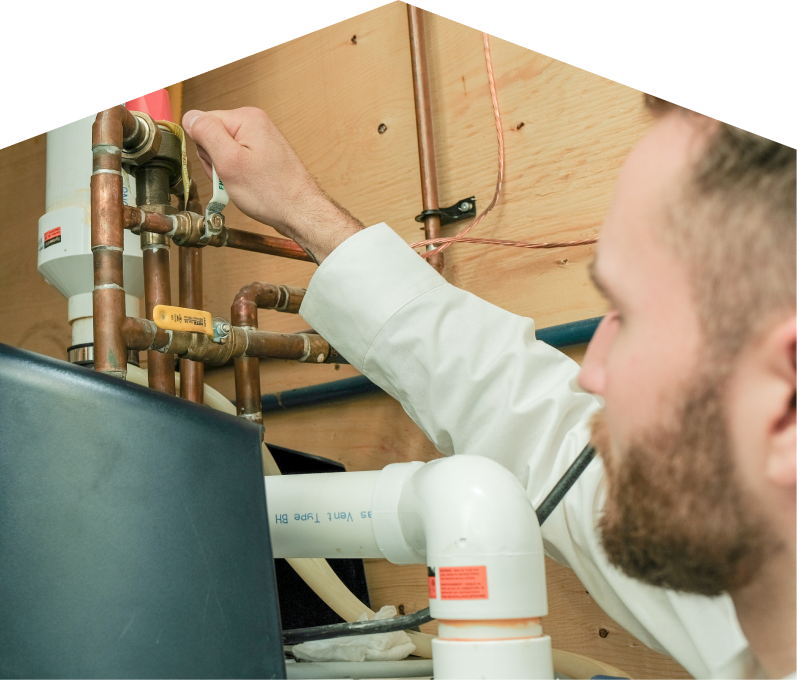 M&K water heater specialist tightens a valve on copper piping during a water heater installation, with exposed wood paneling and wiring in the background. M&K water heater specialist tightens a valve on copper piping during a water heater installation, with exposed wood paneling and wiring in the background.