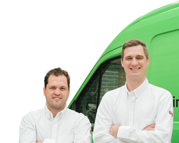 Two M&K technicians smiling in front of their green service van, wearing issued jackets during an on-site consultation.